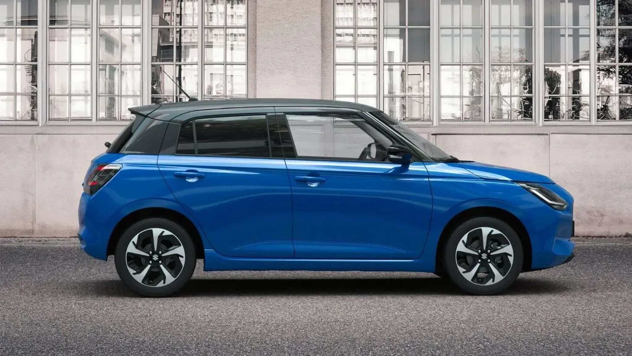 Der neue Suzuki swift in blau mit Anthrazitfarbe im Dach in der Seitenansicht vor einem Lagergebäude